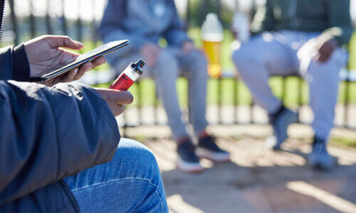 Bild där man ser en person sitta med en mobiltelefon i ena handen och en e-cigarette i den andra. Bilden symboliserar en tonåring som vejpar, det vill säga tonårsvejp.